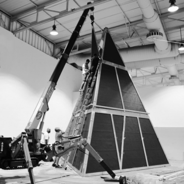 Black and white photograph of a spider crane lifting and positioning a panel at a tall and of monumental dimensions tetrahedral pyramid which is part of an art installation by multimedia artist Pavlina Vagioni at the Athens School of Fine Arts in Greece. Three people are assisting the setup.
