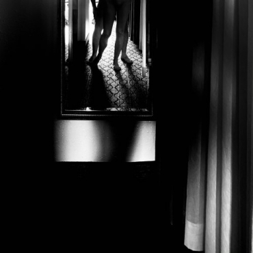 Black and white photo with intense chiaroscuro. At the top, a mirror with a reflection of four legs. A glimpse of a curtain on the right. Black background.