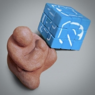 A clay figurine of a seated woman holding a baby rendered in a spherical and minimal manner. Next to it a Rubik's cube with a uniform light-blue background color and white linear shapes on top.