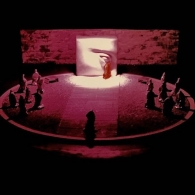 scenographic maquette of the ancient theater of Epidaurus on a scale of 1:100, depicting a circular stage with miniature aluminum foil actors. The focal point of the setting is a sculptured wall with an eye where a red-caped actor stands beneath it creating the illusion of a red tear. The lighting of the stage is centered around this wall.