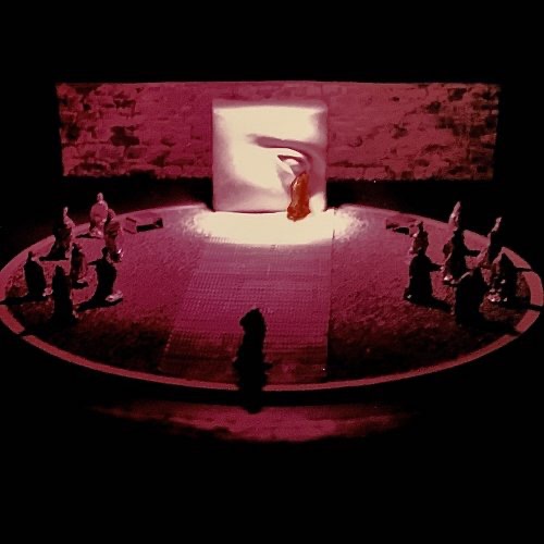 scenographic maquette of the ancient theater of Epidaurus on a scale of 1:100, depicting a circular stage with miniature aluminum foil actors. The focal point of the setting is a sculptured wall with an eye where a red-caped actor stands beneath it creating the illusion of a red tear. The lighting of the stage is centered around this wall.