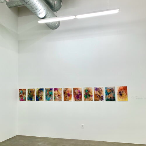 Row of small, colorful abstract paintings mounted in a straight line along a white gallery wall under a fluorescent light and exposed ducts.
