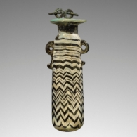 2YX4MW4 Alabastron, 6th-4th century BC. Blue Perfume Flask with an Applicator. 
Additional Info: Core-formed, opaque turquoise glass alabastron decorated with dark blue (cobalt) vertically combed zig-zag pattern in dark blue (cobalt). Spiraling threads of light blue are above the main pattern and decorate the lip edge. Trails of molten blue glass form scroll handles on either side of the upper body. The vessel still has a small section of a bronze chain attached to the mouth.