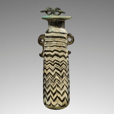 2YX4MW4 Alabastron, 6th-4th century BC. Blue Perfume Flask with an Applicator. 
Additional Info: Core-formed, opaque turquoise glass alabastron decorated with dark blue (cobalt) vertically combed zig-zag pattern in dark blue (cobalt). Spiraling threads of light blue are above the main pattern and decorate the lip edge. Trails of molten blue glass form scroll handles on either side of the upper body. The vessel still has a small section of a bronze chain attached to the mouth.