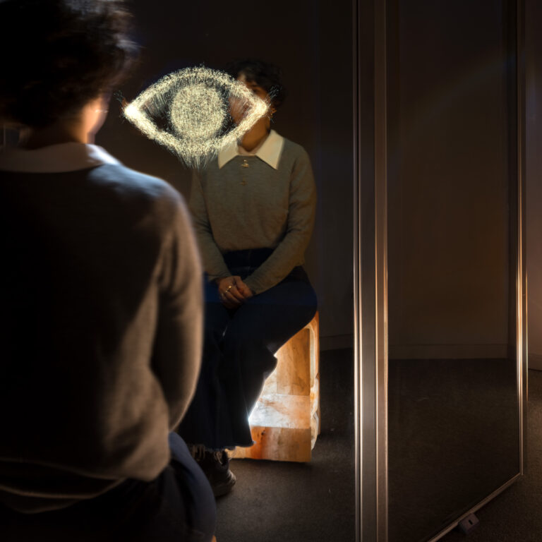 Two people in a dark room; a glowing chalk drawing of an eye on the glass in front of one person, while another person is seen from behind in the foreground.