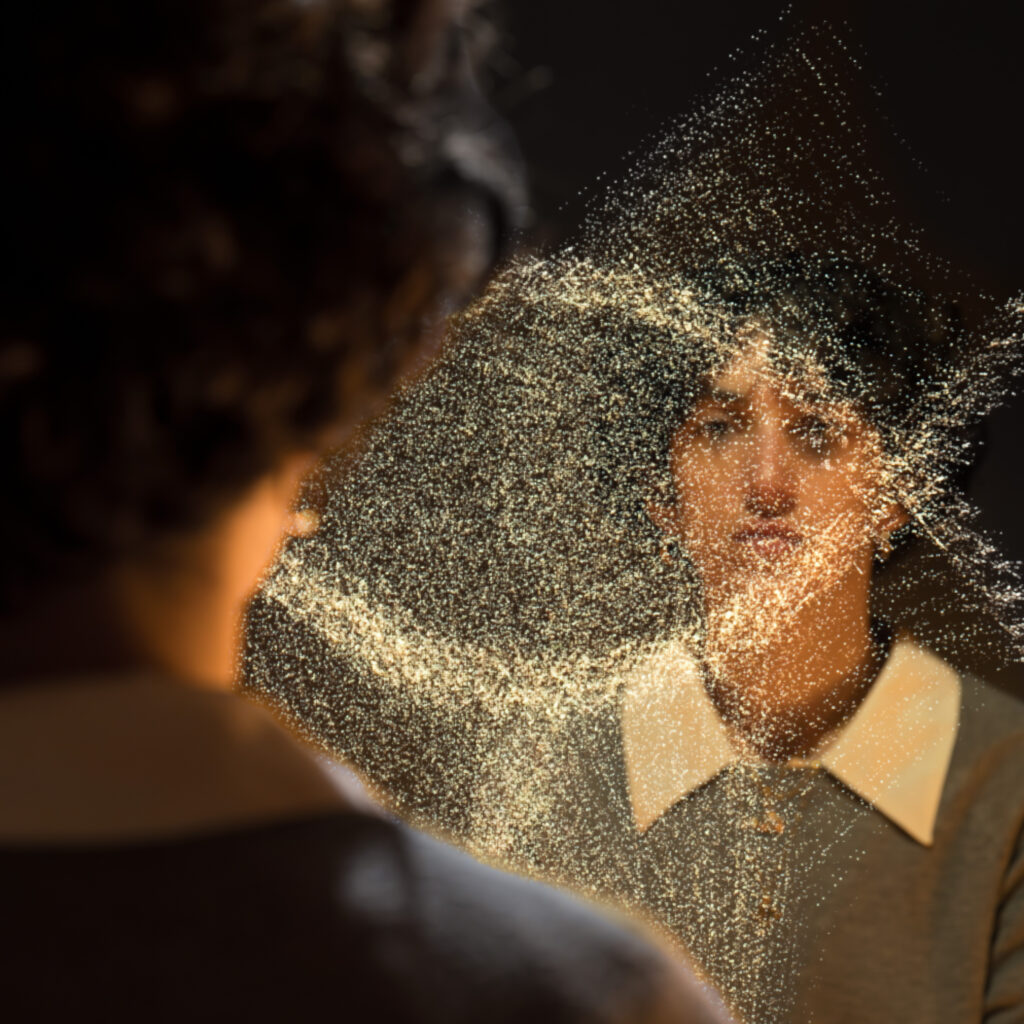 Close-up of a person with golden glitter dusting their face and surroundings in a dark background, creating a sparkling, festive effect. Nearby blurred silhouette suggests another person.