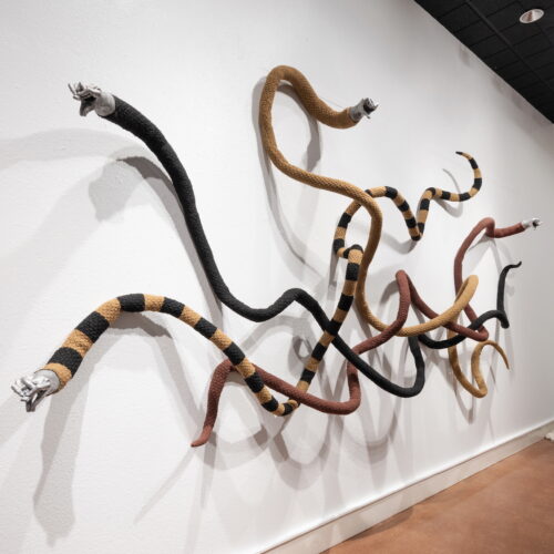 Installation of several long striped rope sculptures twisting along a white gallery wall.