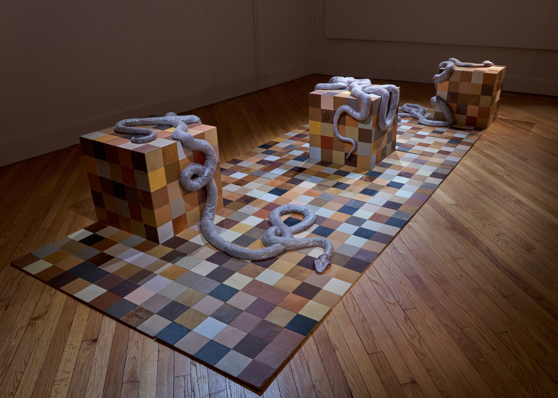 Art installation featuring multicolored tiled blocks connected by a gray rope snaking over a checkered floor in a gallery corner.