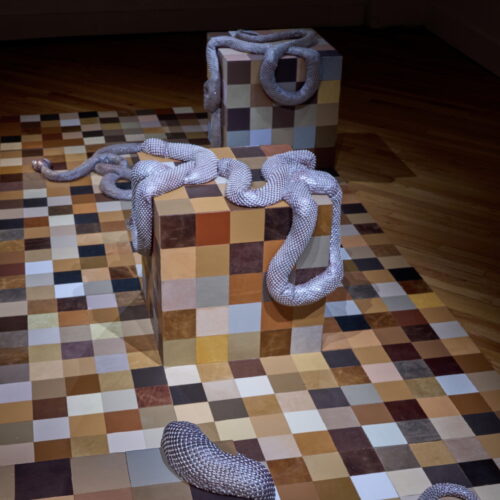 Art installation with large checkered cubes and a silver snakes sculpture weaving around and over the blocks on a multicolored tiled floor.