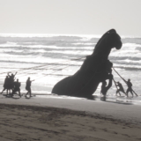 Group of people on a beach pulling ropes attached to a large dark horse sculpture by the water's edge.