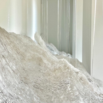 Abstract sculpture featuring a white, textured surface resembling mountains or waves, with thin vertical glass or acrylic rods hanging above/around it to create a reflective, airy effect.