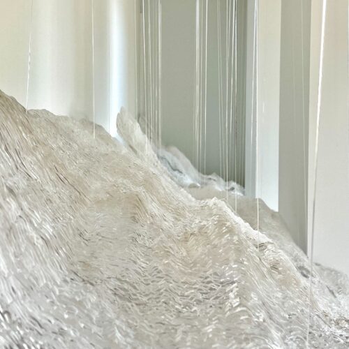 Abstract sculpture featuring a white, textured surface resembling mountains or waves, with thin vertical glass or acrylic rods hanging above/around it to create a reflective, airy effect.