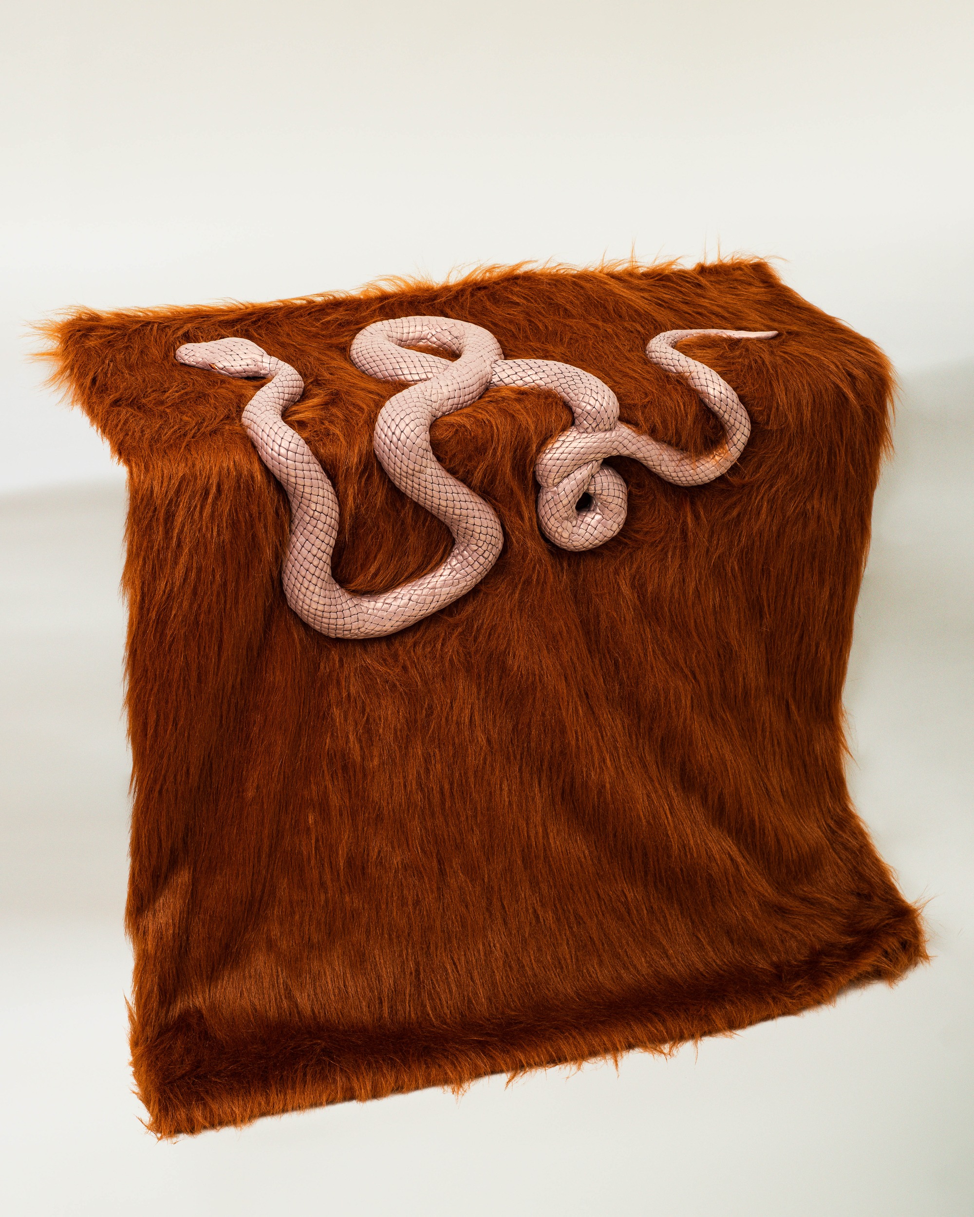 Pink patterned snake curled on a brown faux fur surface, likely an art piece or sculpture.