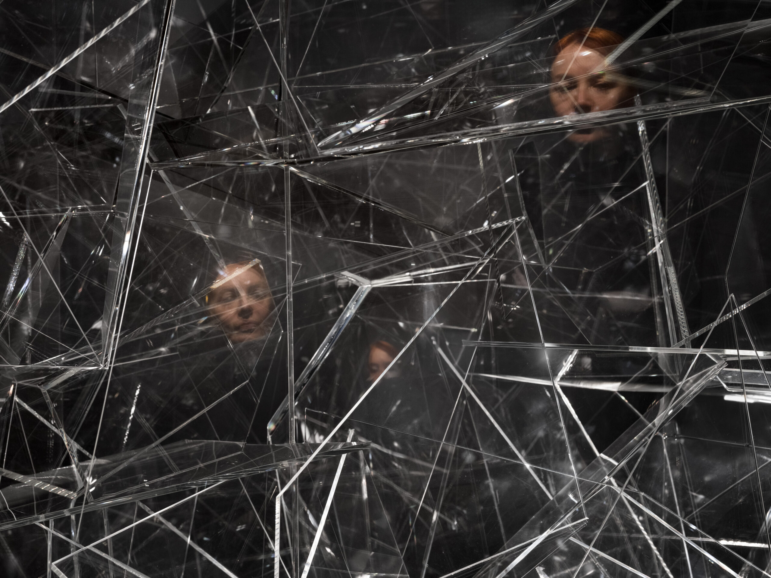 Abstract installation of crisscrossing glass rods reflecting light, with faint human reflections visible in the surfaces.