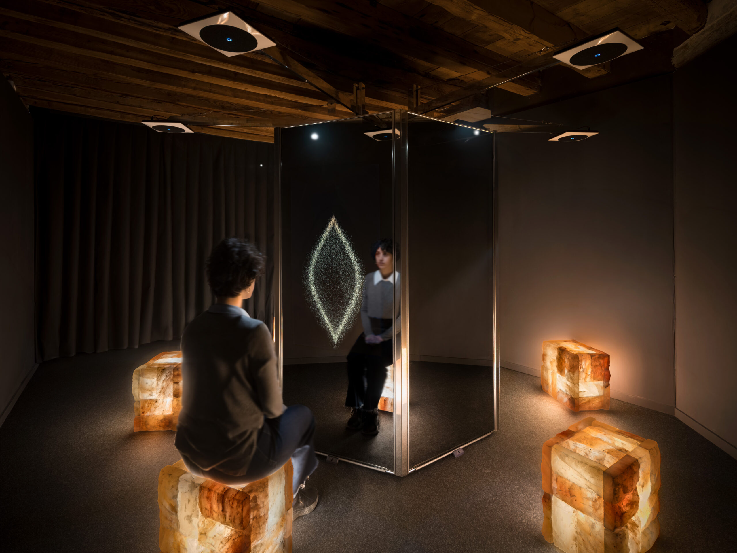 Dimly lit gallery with mirrored panels forming a corner, brick-like blocks on the floor, and a person observing reflective wall art in the corner.
