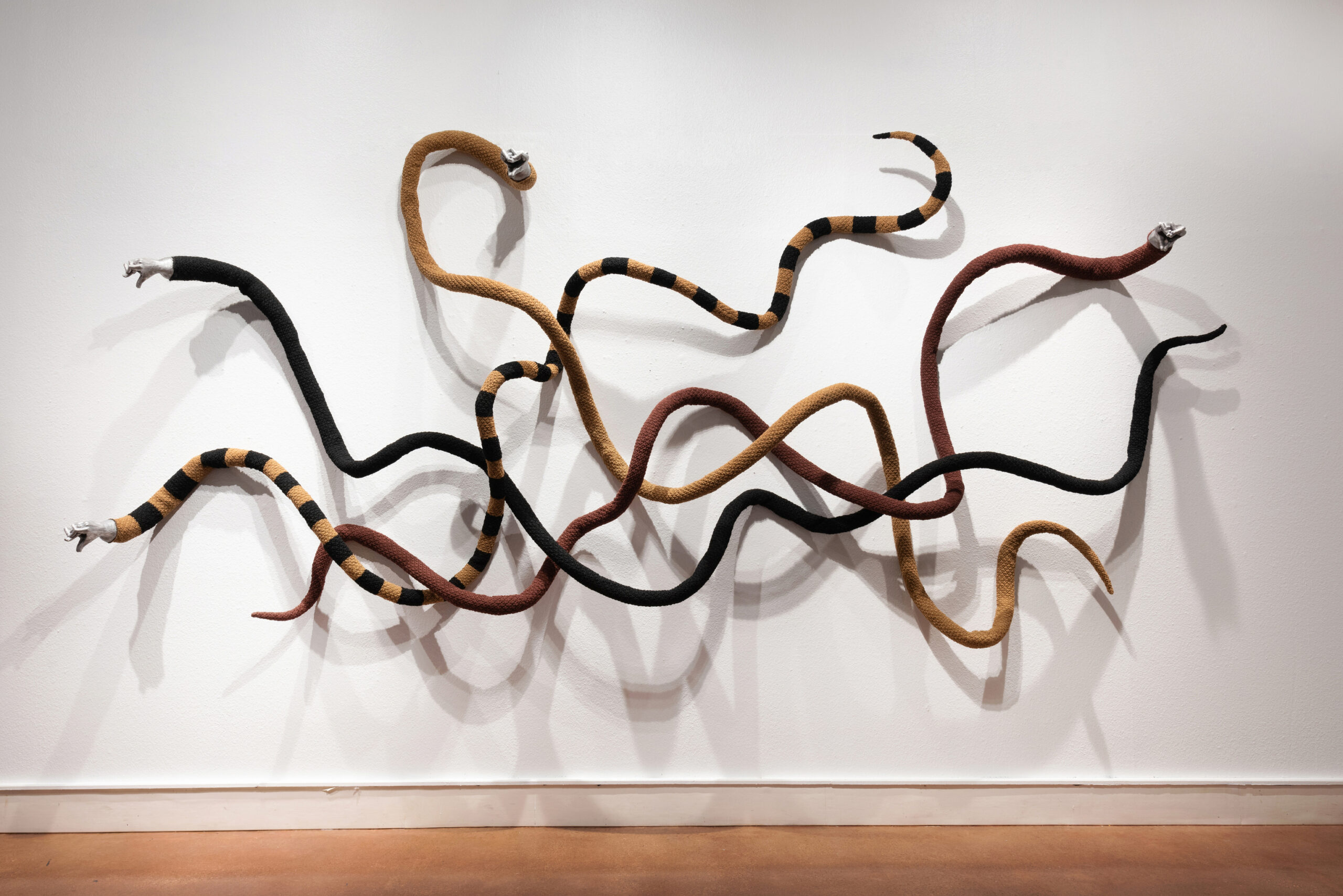 Abstract sculpture of intertwined striped tubes—orange, black, and brown—mounted on a white gallery wall.